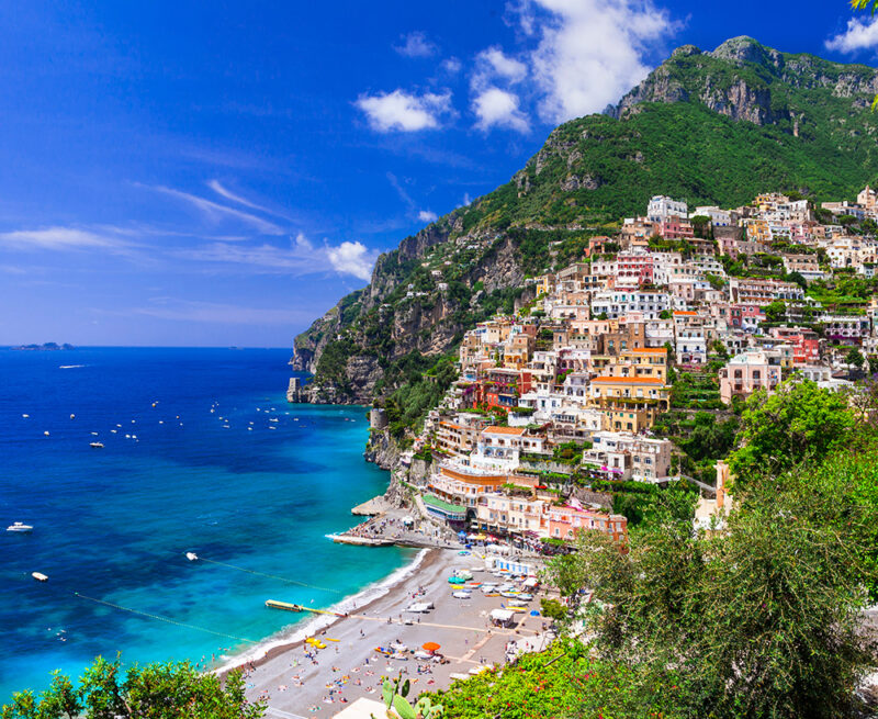 The iconic town of Positano features colorful houses stacked on a lush mountain above a crowded beach on luxury Amalfi Coast trips.