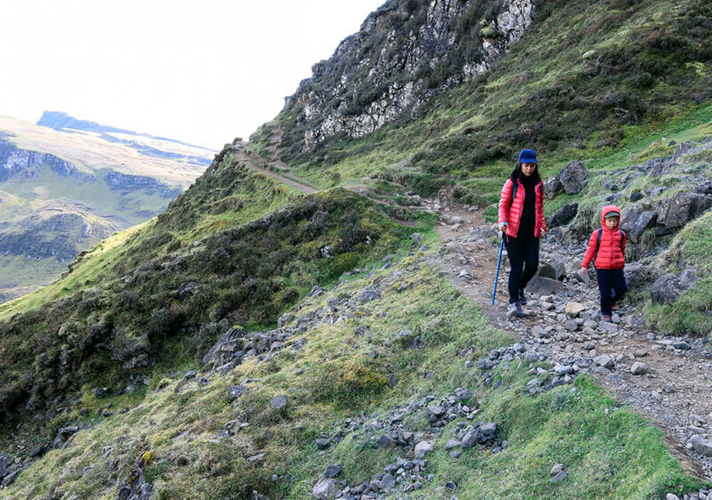 A woman and a small child in red jackets hiking on a steep, rocky mountain trail under a bright sky.