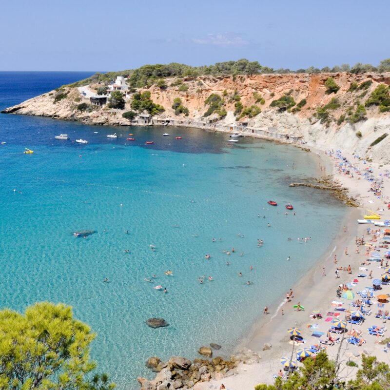 Pine-clad red cliffs curving around the turquoise cove of Cala d’Hort in Ibiza.