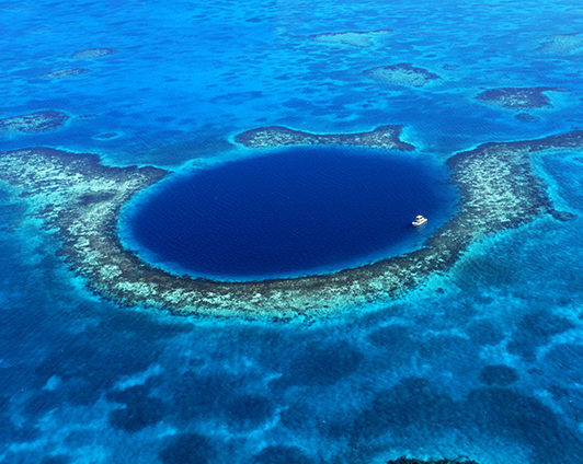 An aerial shot of a large, circular dark blue underwater sinkhole surrounded by turquoise coral reef and a small white boat.