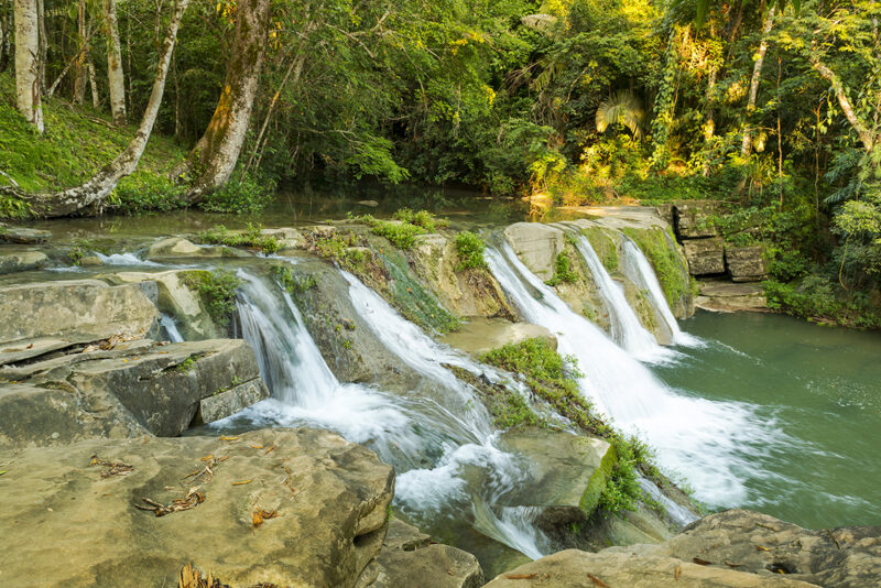 A scenic tropical waterfall flowing into a river within a lush green forest on a sunny day.