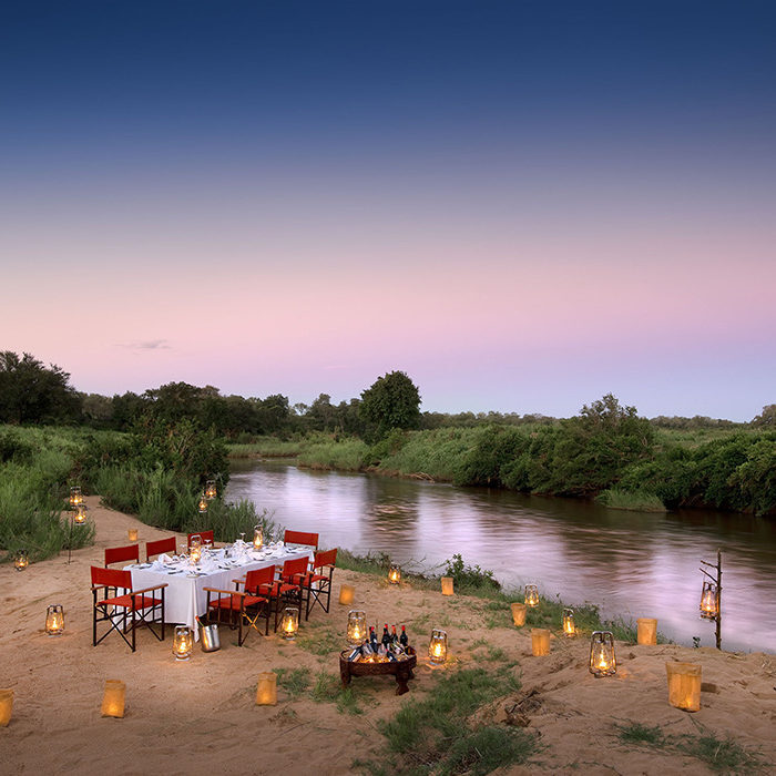 A romantic dinner table with red chairs by a river during luxury Africa honeymoon trips.