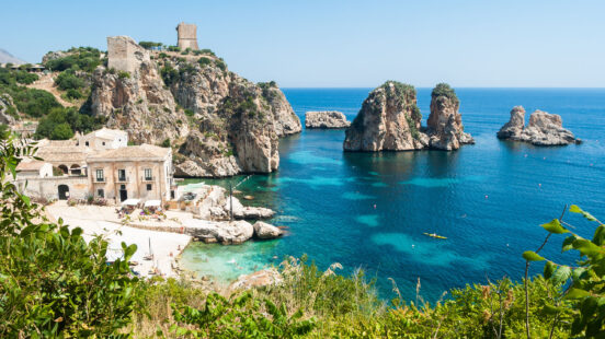 A coastal village with historic stone architecture nestled beside rocky cliffs and sea stacks in the bright blue ocean.