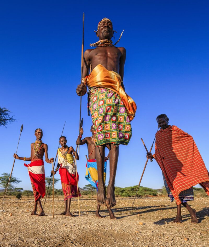 Maasai men in traditional red and patterned clothing performing a jumping dance on luxury Maasai Mara trips.