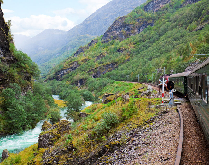 A green train winding around a curved track next to a rushing river in Norway