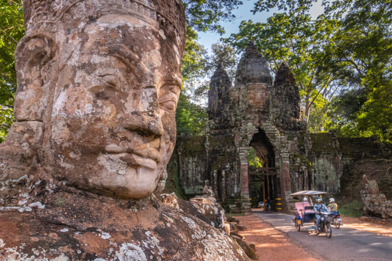 Close-up of a colossal, weathered stone face statue with the ornate, dark temple gate of Angkor Thom in the background.