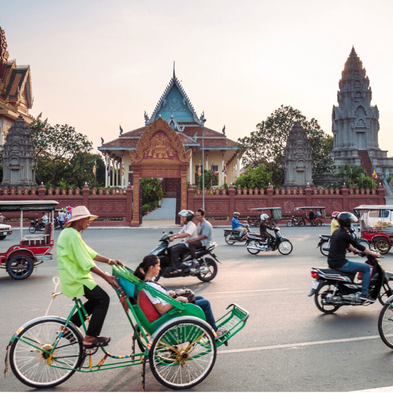 Busy street scene with motorbikes and a cyclo carrying passengers past ornate temple buildings in a Cambodian city.