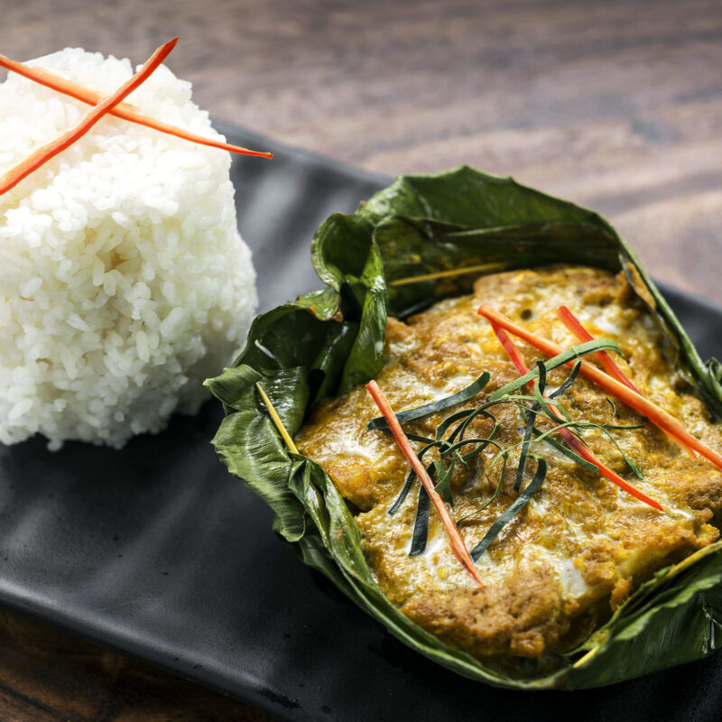 Traditional Cambodian Fish Amok, a yellow fish curry in a banana leaf, next to a tall, neat mound of white rice.