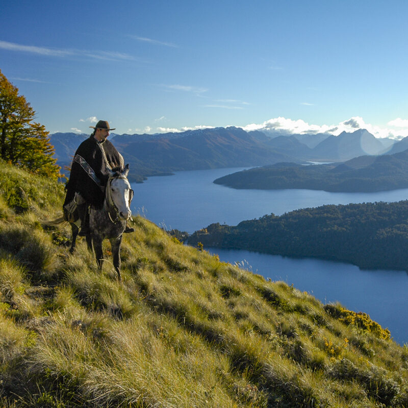 A person riding a horse on a high grassy path above a blue lake