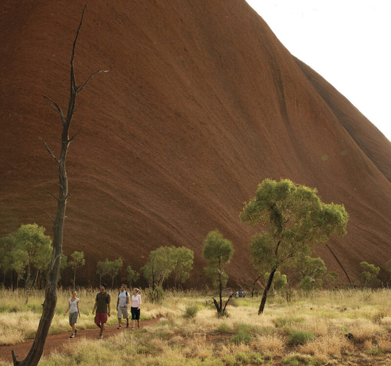 Travellers walk along the base trail beneath the towering sandstone walls of Uluru, Australia.