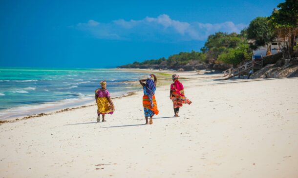 Three women in colorful dresses walk away from the camera on a wide, white sand beach bordering turquoise water.