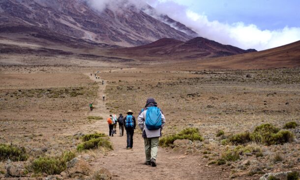 Hikers with backpacks walk along a long, winding dirt path on a rocky mountain under a cloudy sky.