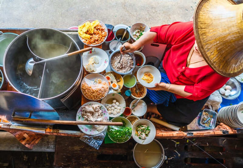 A street food vendor in a conical hat prepares bowls of noodles and various ingredients at a floating market. luxury food and wine tours.