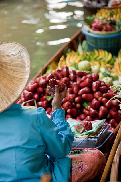 A vendor in a blue shirt holds a red fruit in a boat filled with tropical fruit. luxury food and wine tours