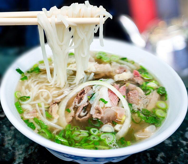 Chopsticks lifting noodles from a bowl of Pho soup with meat, onion, and herbs. luxury food and wine vacations.