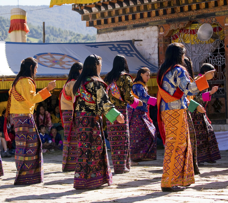 A group of women in vibrant, patterned traditional Bhutanese clothing perform a coordinated dance outdoors.