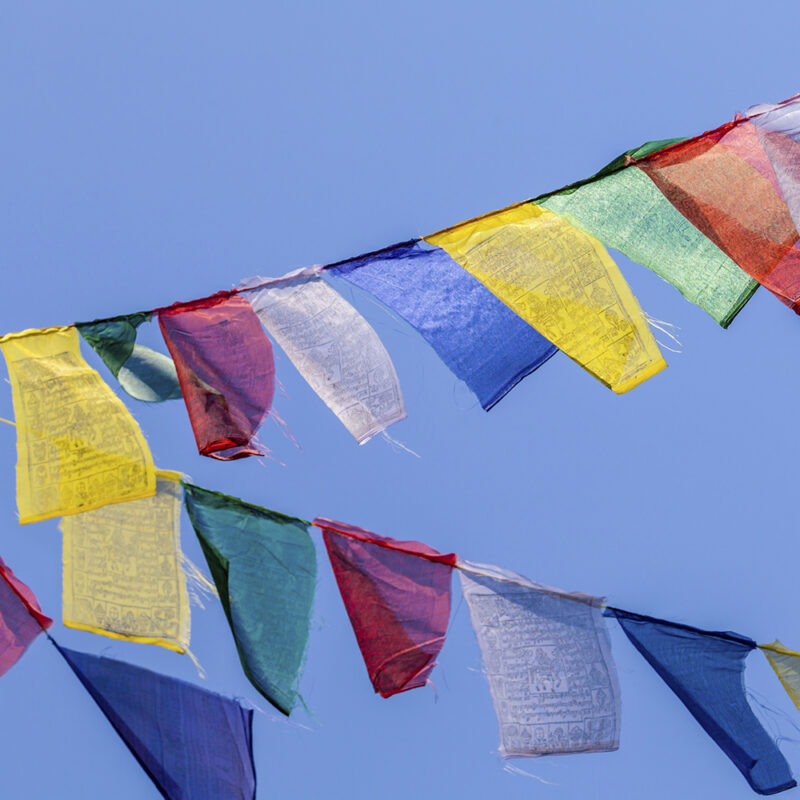 Multiple rows of colorful rectangular prayer flags strung across a vivid blue sky.