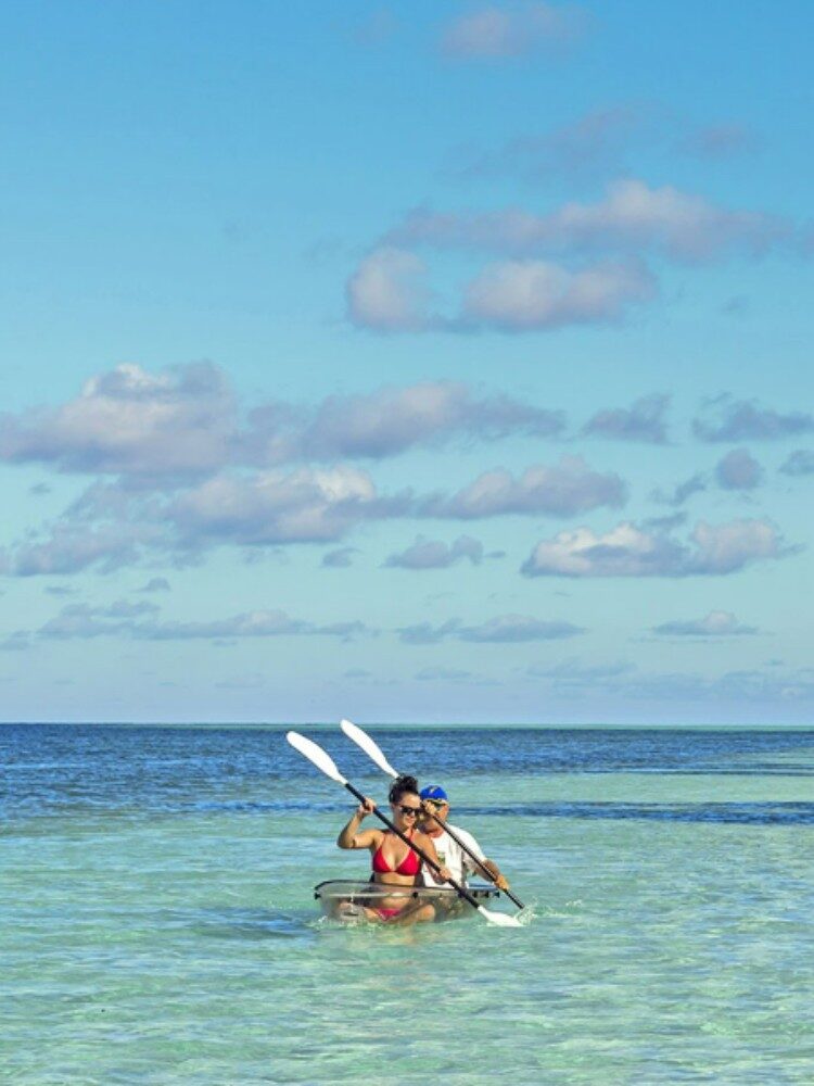 Two kayakers paddling in a clear-bottom kayak through shallow Seychelles waters.