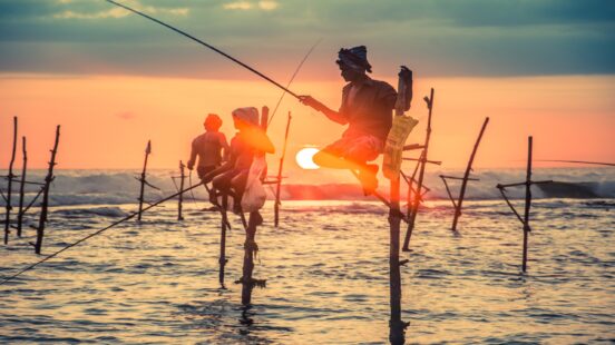 Multiple fishermen sitting on wooden stilts in the ocean waves at sunset.