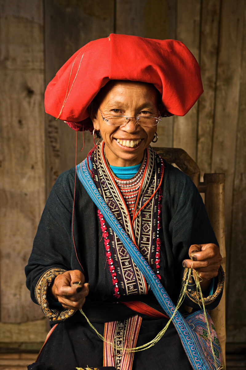 Woman in a large red headdress and traditional embroidered clothing sewing for luxury culture tours.