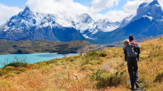 A man carries a child on his back while hiking a mountain trail near a blue lake during luxury Family vacations.