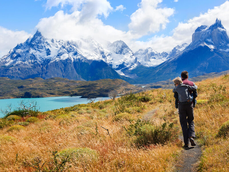 A man carries a child on his back while hiking a mountain trail near a blue lake during luxury Family vacations.