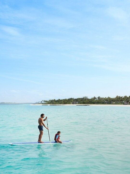 Two people on a single paddleboard in clear blue water near a white sand beach during luxury Family vacations.