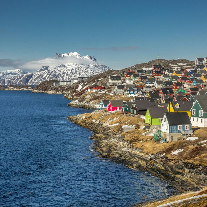 Luxury European Tours - The town of Nuuk, Greenland, with rainbow painted traditional hut-style houses on a rocky shoreline, with snowy mountains in the background