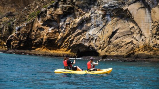 Two people paddling a yellow tandem kayak in the ocean next to a large, textured rocky cliff face.