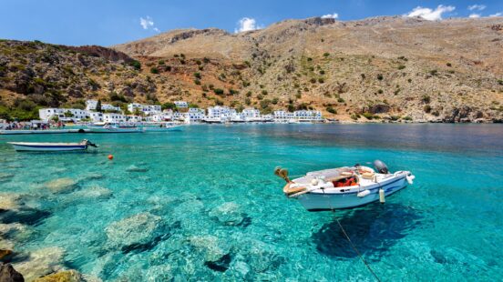 Little boats floating on crystal clear water in a bay next to a small Greek village with white sugar-cube buildings