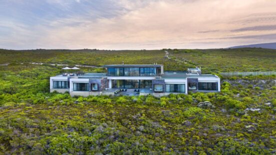 Aerial view of a modern rectangular luxury house surrounded by dense green vegetation and rolling hills.