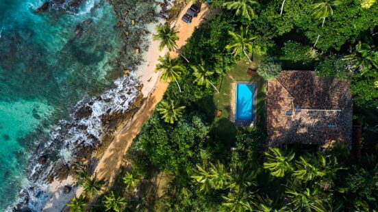 High-angle view of a house with a blue pool and a brown roof located next to a rocky beach.