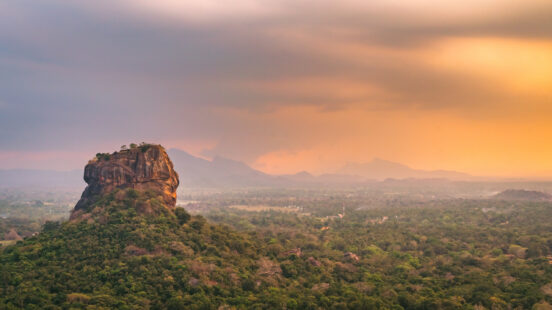 A large, flat-topped rock formation rises above a vast green forest under a warm sunset sky.