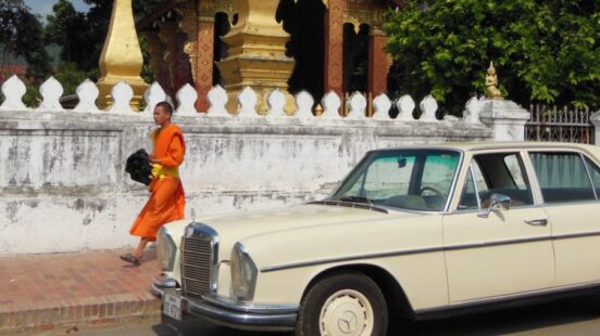 A monk in orange robes walking past a vintage cream Mercedes parked on a street.
