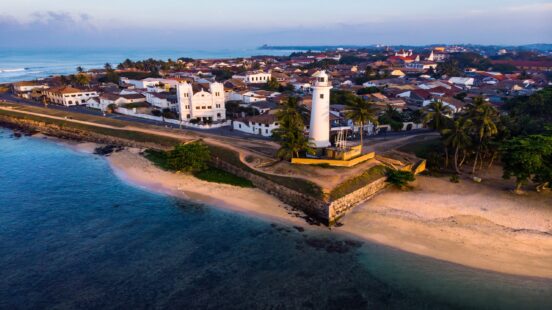 A tall white lighthouse stands on a stone fort wall by the ocean at sunset.