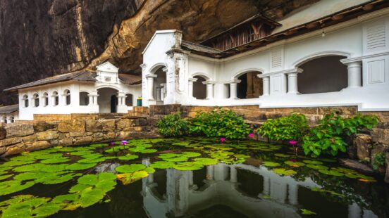 A white temple building built into a rock face with a lily pond in the foreground.