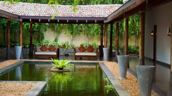 A peaceful courtyard with a rectangular pond reflecting green plants, surrounded by pillared walkways and sofas.