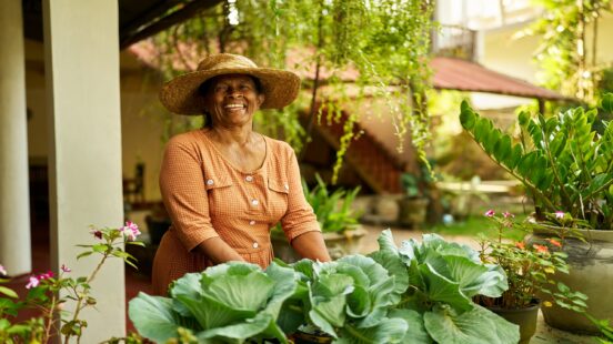 A woman in a wide-brimmed hat smiling behind lush green vegetables in a garden.