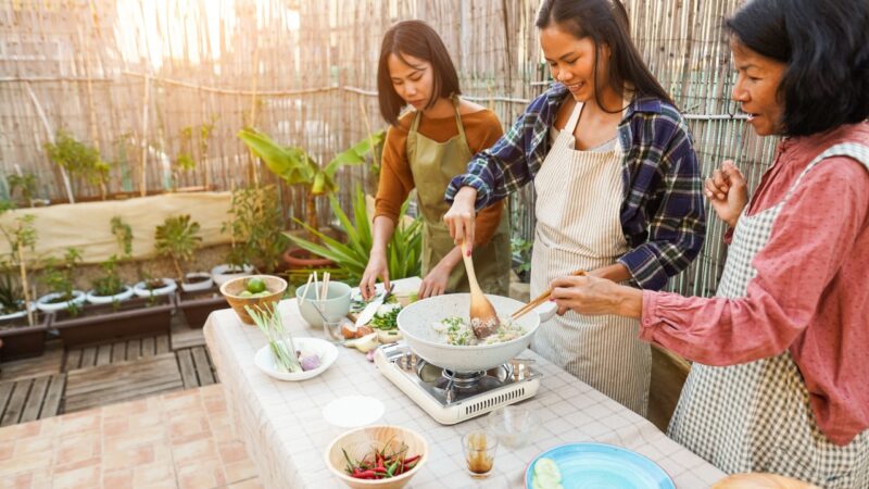 Three women in aprons cooking together at an outdoor table with a wok and various fresh ingredients.