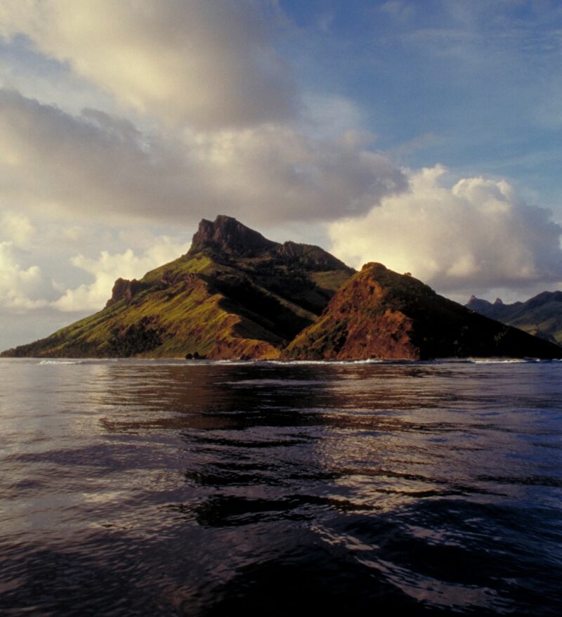Rugged green and brown volcanic mountains on an island surrounded by dark ocean waves for luxury Fiji vacations.