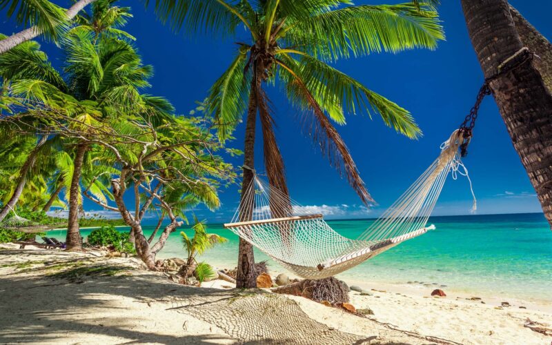 Rope hammock tied to palm trees on a white sand beach overlooking a tropical blue sea for luxury Fiji vacations.