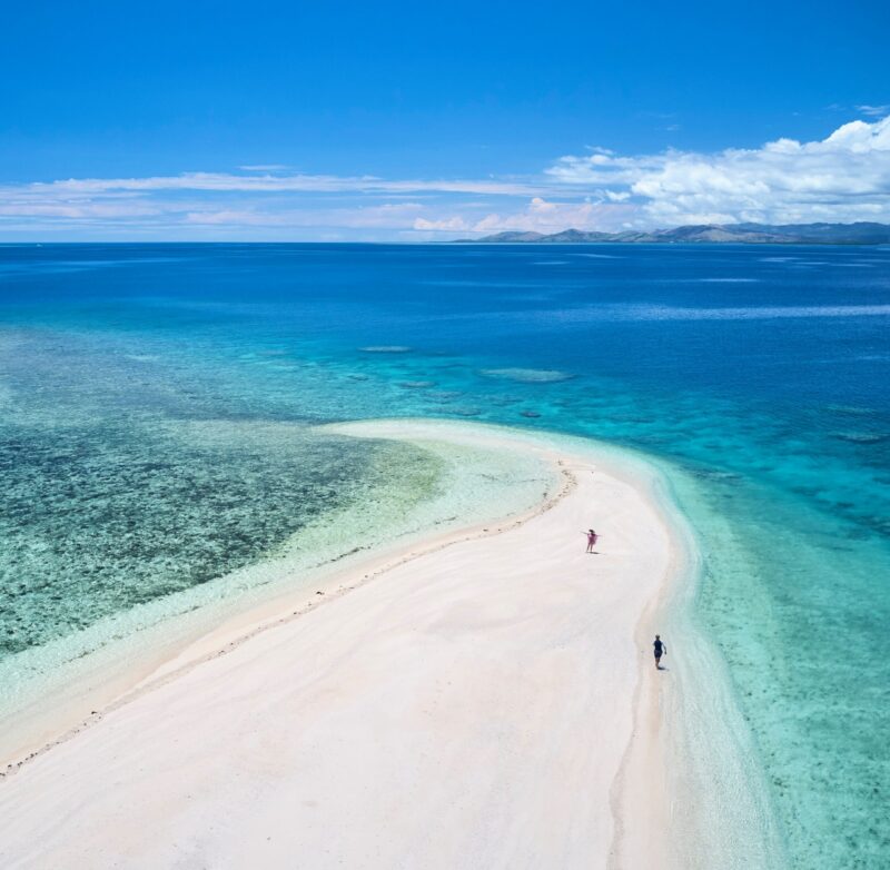 Aerial view of a long white sandbar surrounded by turquoise and deep blue ocean during luxury Fiji trips.
