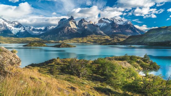 A calm turquoise lake reflects the surrounding mountains and rugged terrain under a blue, cloud-filled sky.
