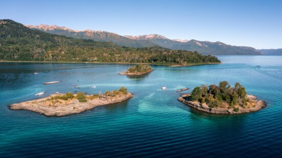 High-angle view of several small islands with green trees in a vast, clear blue lake near distant mountains.