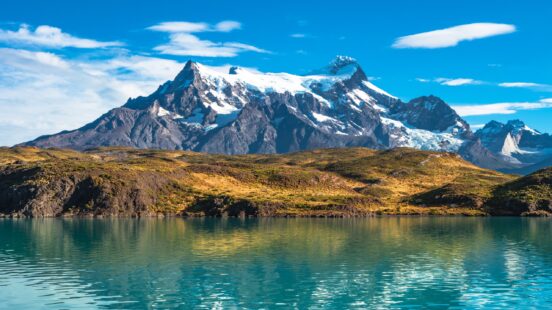 Large snow-capped mountain peaks towering over a turquoise lake with golden grassy hills in the foreground.