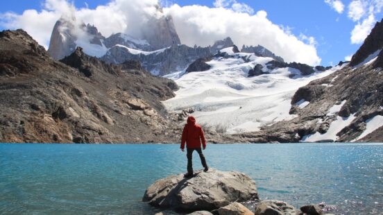 A person in a red jacket stands on a rock by a turquoise glacial lake facing massive snow-capped mountains.