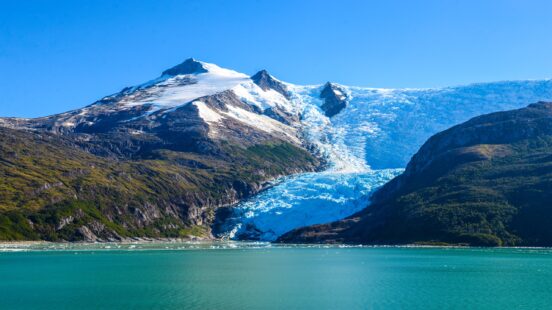 A large blue glacier descends from a mountain peak into a vast lake under a bright blue sky.