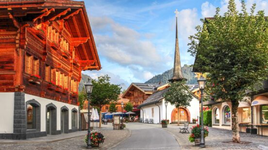 Traditional wooden chalets and a church spire along a paved village street under a cloudy sky.