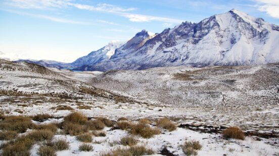 A wide, snowy landscape with low shrubs leading toward massive, snow-capped mountains in the distance.