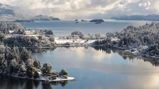 A snowy landscape shows a building on a hill overlooking a peaceful lake with trees dusted in white snow.